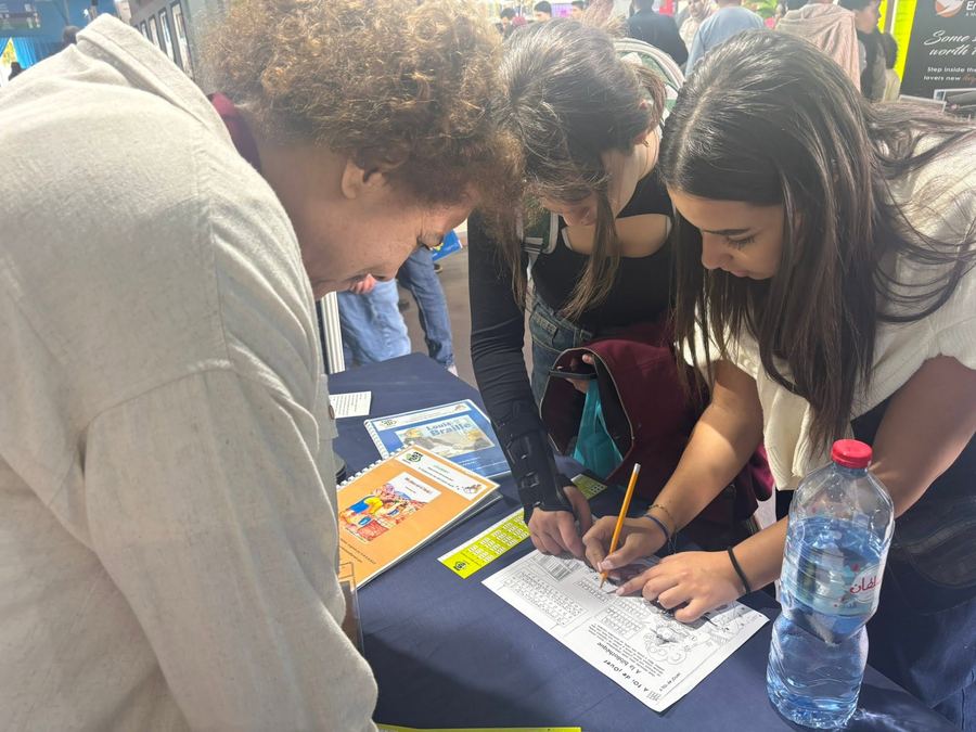 Deux jeunes visiteuses s'exercent à l'écriture braille avec une règle et un poinçon, à côté d'un livre sur Louis Braille