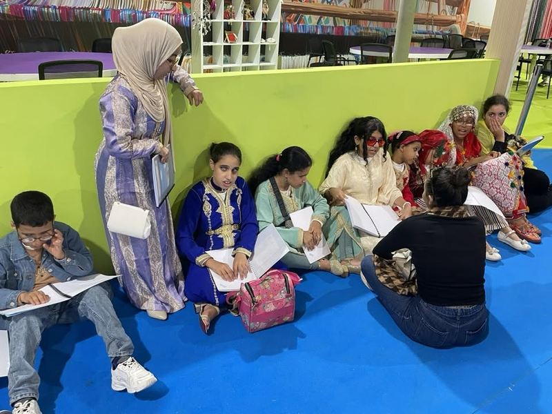 Un groupe d'enfants assis au sol lit des livrets reçus, encadré par une bénévole