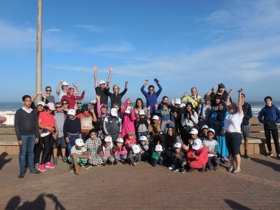 Un grand groupe de bénéficiaires, familles et bénévoles d'AMARDEV pose joyeusement, bras levés, sur la corniche en bord de mer lors d'une sortie de l'association